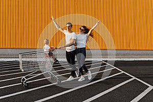 Happy family, husband and wife with child sitting in shopping trolley making purchases against yellow wall of shopping mall,