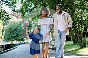 Happy family, joyful time together outdoors. Dark skinned little girl in blue dress, blowing colorful windmill toy