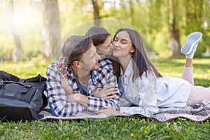 Happy family with kid lying on green grass in park. Mother and father hugging and kissing son.