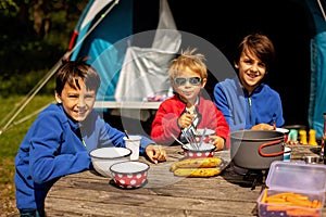 Happy family, kids, boy brothers and a dog, having breakfast in front of pitched tent in the forest, while wild camping in Norway