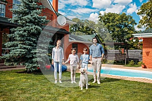 happy family with labrador dog looking at camera while holding hands on backyard of country house