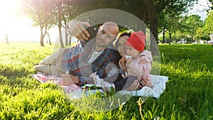 Happy family is laying on the grass and doing selfie with a baby at sunset in the park. Father and mother take pictures