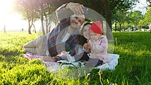 Happy family is laying on the grass and father doing selfie with a baby at sunset in the park on the smartphone