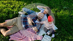 Happy family is laying on the pled and doing selfie with a baby at sunset in the park. Father and mother take pictures