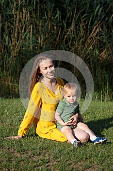 Happy family leisure outdoors. Portrait of smiling young woman holding little toddler son in arms. They are sitting on the green