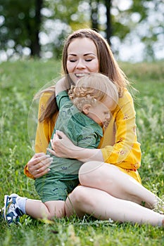 Happy family leisure outdoors. Portrait of smiling young woman holding little toddler son in arms. They are sitting on the green