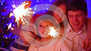 Happy family lit sparklers at the party. In the background, bokeh lights and garlands of Christmas fir