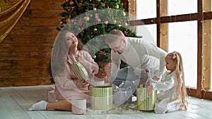 Happy family looking gifts over Christmas tree on holiday morning. Mod, dad and two daughter on floor opening gift boxes