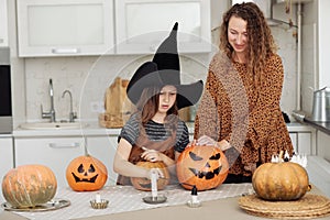 Happy family: mom and daughter celebrate Halloween. Merry child in witch hat at home kitchen. Cheerful kid and parent