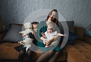 Happy family, mom hugs little daughter sitting at home on the couch. The girl is holding a white fluffy dog in her hands