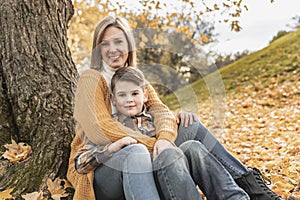 Happy family mother and child boy in the autumn leaf fall in park sit on the bottom of the tree