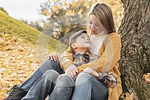 Happy family mother and child boy in the autumn leaf fall in park sit on the bottom of the tree