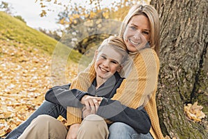 Happy family mother and child boy in the autumn leaf fall in park sit on the bottom of the tree