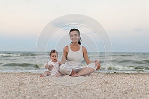 Happy family mother and child daughter doing yoga, meditate in lotus position on beach at sunset