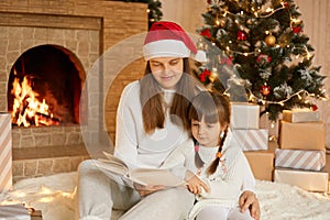 Happy family mother and child daughter reading book on winter evening near fireplace and xmas tree, mommy and female kid sitting