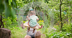 Happy family, mother and child play on wooden swing in garden under tree