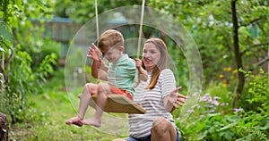 Happy family, mother and child play on wooden swing in garden under tree