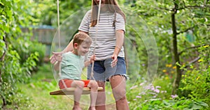 Happy family, mother and child play on wooden swing in garden under tree