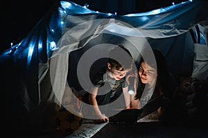 Happy family mother and child son reading a book with a flashlight in a tent at home. family concept