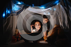 Happy family mother and child son reading a book with a flashlight in a tent at home. family concept