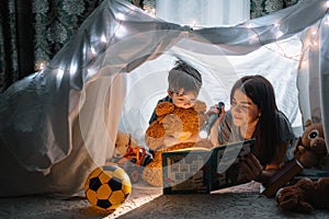 Happy family mother and child son reading a book with a flashlight in a tent at home. family concept