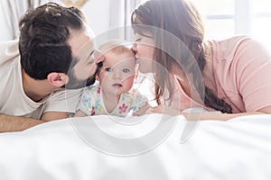 Happy family, mother, father and baby on the white bed giving side kiss