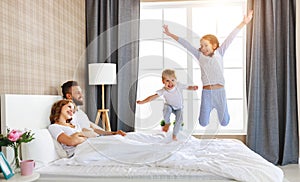 Happy family mother, father and children laughing, playing and jumping in bed   at home