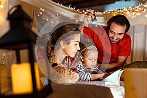 Happy family, mother, father and daughter lying inside hut, tent in room in the evening with Christmas lights and