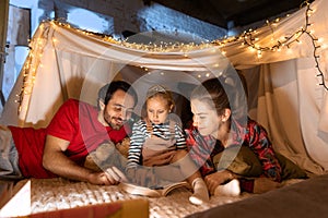 Happy family, mother, father and daughter lying inside self-made hut, tent in room in the evening and reading book