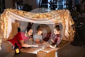 Happy family, mother, father and daughter lying inside self-made hut, tent in room in the evening and reading book