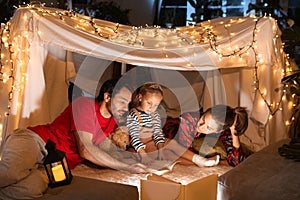 Happy family, mother, father and daughter lying inside self-made hut, tent in room in the evening and reading book