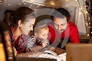 Happy family, mother, father and daughter in pajamas lying inside self-made hut, tent in room in the evening and reading