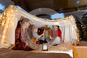 Happy family, mother, father and daughter playing inside hut with lights, tent in room in the evening. Pillows fight