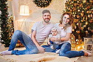 Happy family of mother, father and little baby girl sitting near Christmas tree on the floor