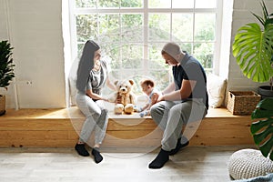 Happy family mother and father playing with a baby at home sitting on the windowsill