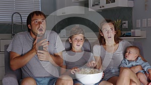 Happy family mother father and teen kids are watching scary movie on TV expressing fear and talking sitting on couch at