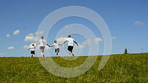 Happy family mother, father and two little sons run in a green meadow holding hands on a background of blue sky and clouds