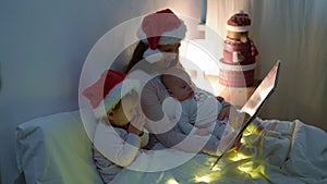 happy family mother in red santa hat two children babies little baby siblings Christmas night in bed pajamas have fun