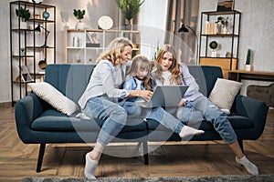 Happy family of mother and two cute daughters sitting together on comfortable couch, using laptop