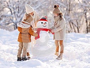 Happy family mother and two little kids making snowman together in winter park outdoors