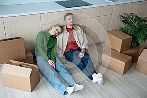 Happy family moving into new apartment, sitting on floor, resting, going to unpack cardboard boxes