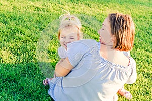 Happy family on nature. Mom and toddler baby daughter relaxing, hugging, laughing and have fun on green grass meadow at evening su