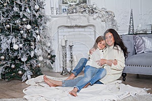 Happy family on New Year Eve. Beautiful mother and cute daughter in white cozy sweaters have fun and hug under a decorated