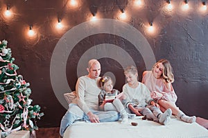 Happy family in the new year`s interior sitting on the bed and reading a book. They`re smiling. Next is a Christmas tree