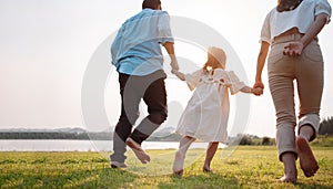 Happy family in the park sunset light. family on weekend running together in the meadow with river Parents hold the child hands.