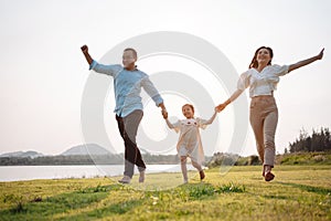 Happy family in the park sunset light. family on weekend running together in the meadow with river Parents hold the child hands.