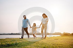 Happy family in the park sunset light. family on weekend running together in the meadow with river Parents hold the child hands.