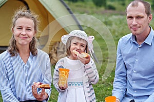 Happy family on picnic at camping. Mother, father and daughter eating near a tent in meadow or park