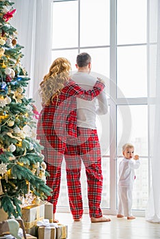 Happy family in plaid pajamas near the big window in the living room with a Christmas tree. Mother, father and little son waiting