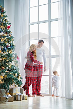 Happy family in plaid pajamas near the big window in the living room with a Christmas tree. Mother, father and little son waiting